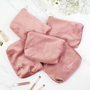 Pu&ograve; includere: Cinque pochette con cerniera in velluto rosa con lettere dorate. Le pochette sono personalizzate con i nomi "Natalie", "Heather", "Olivia", "Vicki" e "Amelia".