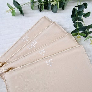 May include: Four beige zippered pouches with gold-colored zippers and tassels. Each pouch has initials printed in white, with a small heart symbol below. The pouches are arranged in a fan shape, with greenery in the background.
