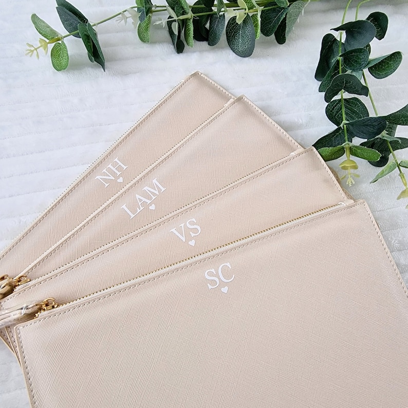 May include: Four beige zippered pouches with gold-colored zippers and tassels. Each pouch has initials printed in white, with a small heart symbol below. The pouches are arranged in a fan shape, with greenery in the background.