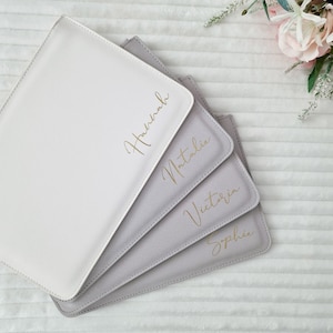 May include: Four personalized, square-shaped, light-colored placemats. The placemats are in shades of white and gray, with gold script names: Hannah, Natalie, Victoria, and Sophie. The placemats are arranged in a fan shape, with a flower arrangement in the background.