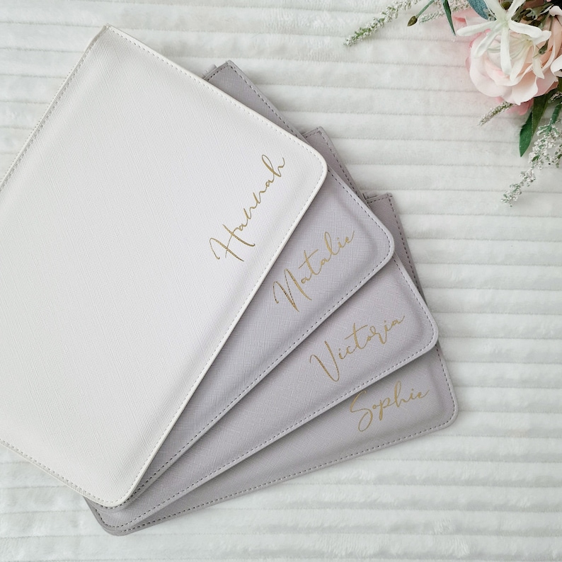 May include: Four personalized, square-shaped, light-colored placemats. The placemats are in shades of white and gray, with gold script names: Hannah, Natalie, Victoria, and Sophie. The placemats are arranged in a fan shape, with a flower arrangement in the background.