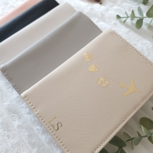 May include: A set of four passport holders in different colors, including beige, gray, black, and pink. The beige passport holder has a gold design of a camera, heart, globe, and airplane. The passport holder is personalized with the initials "LS".