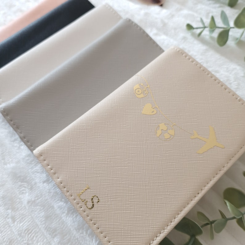 May include: A set of four passport holders in different colors, including beige, gray, black, and pink. The beige passport holder has a gold design of a camera, heart, globe, and airplane. The passport holder is personalized with the initials "LS".