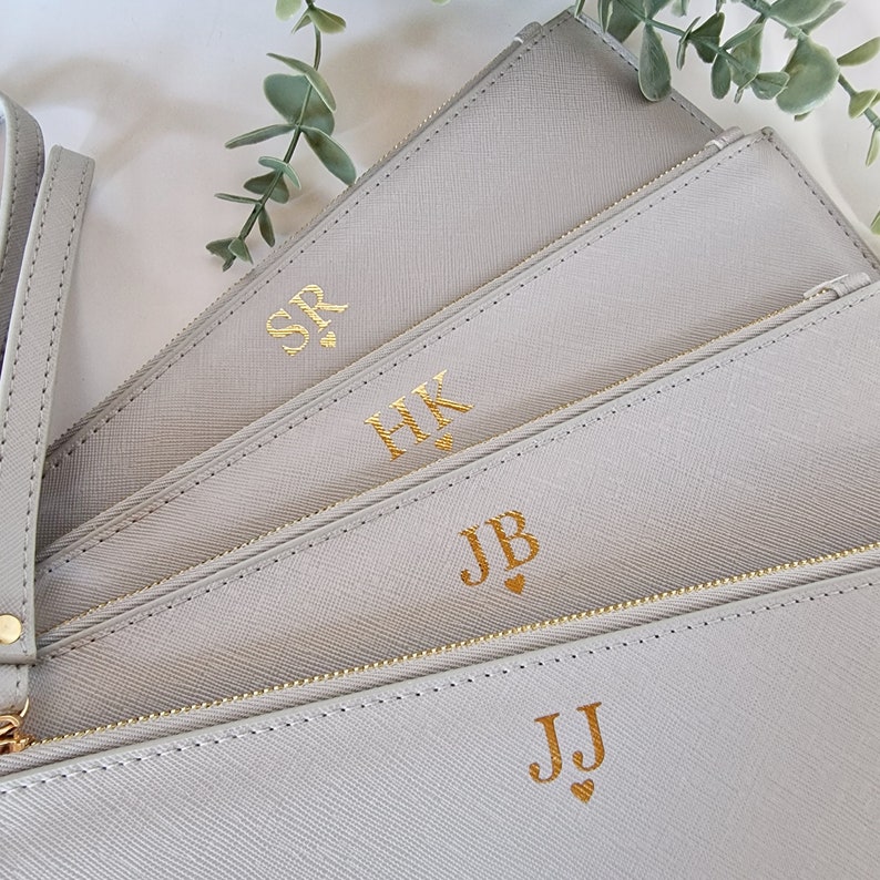 May include: Four light gray zippered pouches with gold-colored initials. The pouches are arranged in a fan shape, with a wrist strap visible on the left. The initials are SR, HK, JB, and JJ, each with a small heart.