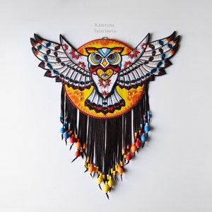 Op de afbeelding: Kleurrijke uil wanddecoratie met uitgespreide vleugels. De uil is afgebeeld met ingewikkelde details in geel, rood, blauw en wit. Zwarte franjes met kralenaccenten hangen eronder. Het kunstwerk is geplaatst tegen een witte achtergrond.