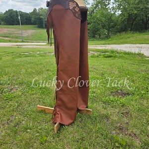May include: Brown leather chaps with fringe and a tooled floral design. The chaps are displayed on a wooden stand against a grassy background.