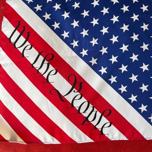 May include: A patriotic flag design featuring the American flag with red and white stripes, a blue field of white stars, and the words "We the People" in black script. The flag is displayed on a red surface.