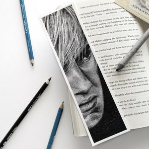 Puede incluir: Marcap&aacute;ginas en blanco y negro con un dibujo a l&aacute;piz de un rostro, parcialmente dentro de un libro. L&aacute;pices azules y negros est&aacute;n dispersos, resaltando el tema art&iacute;stico.
