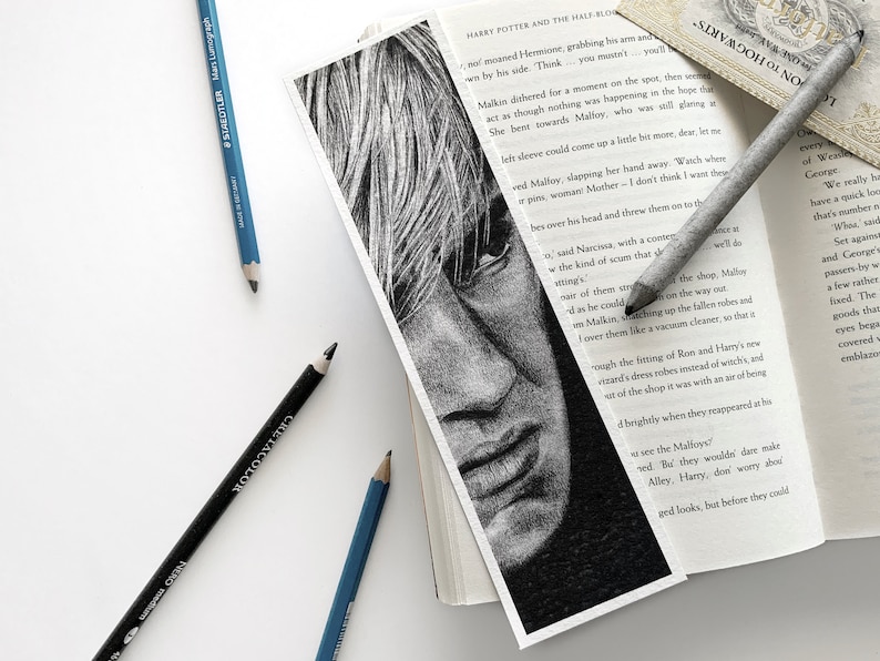 Puede incluir: Marcap&aacute;ginas en blanco y negro con un dibujo a l&aacute;piz de un rostro, parcialmente dentro de un libro. L&aacute;pices azules y negros est&aacute;n dispersos, resaltando el tema art&iacute;stico.