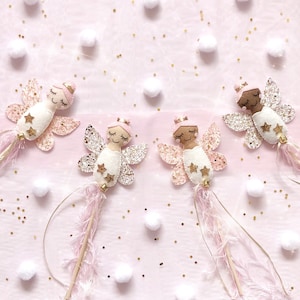 Peut inclure: Quatre baguettes de f&eacute;e avec des t&ecirc;tes de poup&eacute;e en feutrine aux yeux ferm&eacute;s et chignons roses, fix&eacute;es sur des b&acirc;tons en bois. Chaque baguette a des ailes paillet&eacute;es, deux petites &eacute;toiles dor&eacute;es, une clochette dor&eacute;e et des rubans roses.