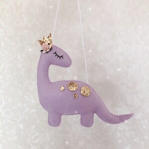 May include: A lavender felt dinosaur ornament hanging from a white string. The dinosaur features a gold glitter crown, a small pink paper flower, embroidered closed eyes, and several gold glitter circular spots on its back.