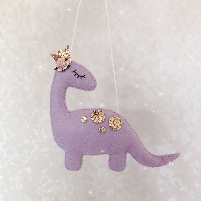 May include: A lavender felt dinosaur ornament hanging from a white string. The dinosaur features a gold glitter crown, a small pink paper flower, embroidered closed eyes, and several gold glitter circular spots on its back.