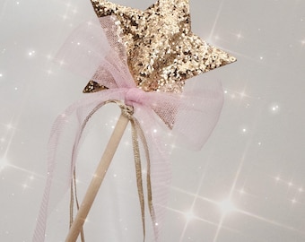 May include: A whimsical star-shaped wand with a gold glitter finish. The star is attached to a light wooden dowel with a pink tulle bow and gold ribbons. The wand is a magical accessory.