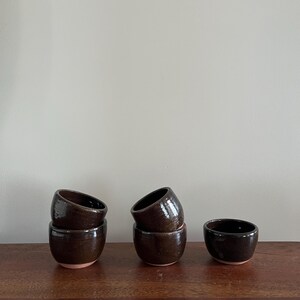 May include: Several small, dark brown ceramic cups with a glossy finish are arranged on a wooden surface. The cups are stacked in pairs. The interior of the cups is a darker shade of brown.
