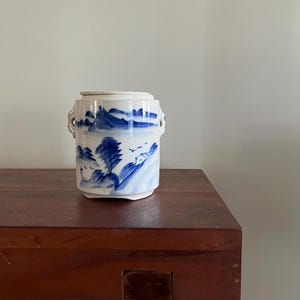 Puede incluir: Un jarrón cilíndrico de cerámica con tapa, con un diseño de paisaje azul y blanco. El jarrón tiene dos asas pequeñas y se exhibe sobre una mesa de madera oscura. El diseño incluye montañas, árboles y agua, creando una estética serena.