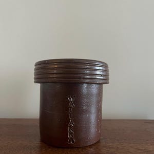 Puede incluir: Un tarro de cerámica marrón con tapa acanalada. El tarro tiene forma cilíndrica con la palabra "WALLAROO" inscrita verticalmente en el lateral. El tarro está sobre una superficie de madera.