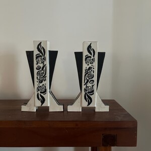 Könnte beinhalten: Ein Paar dekorative Buchstützen. Jede Buchstütze hat eine weiße, rechteckige Säule mit einem schwarzen Blumendesign und schwarzen dreieckigen Stützen. Die Buchstützen stehen auf einem dunkelbraunen Holztisch.