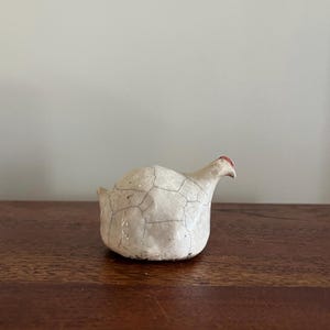 May include: A small, ceramic hen figurine with a white body and a red comb. The hen has a cracked, textured surface and is perched on a wooden surface. The figurine is a decorative item.