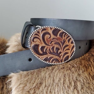 Puede incluir: Un cintur&oacute;n de cuero gris con una hebilla ovalada con un dise&ntilde;o floral marr&oacute;n y beige. La hebilla tiene un borde plateado en forma de cuerda. El cintur&oacute;n descansa sobre una superficie marr&oacute;n y peluda.