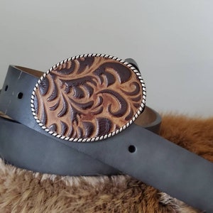 Puede incluir: Un cintur&oacute;n de cuero negro con una hebilla ovalada de cuero tallado marr&oacute;n y beige. La hebilla tiene un ribete de cuerda plateada y un dise&ntilde;o floral detallado. El cintur&oacute;n est&aacute; doblado.