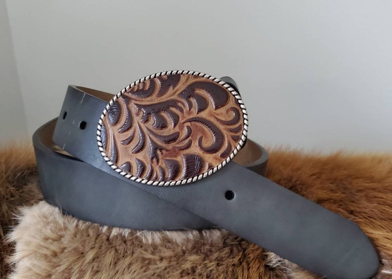 Puede incluir: Un cintur&oacute;n de cuero negro con una hebilla ovalada de cuero tallado marr&oacute;n y beige. La hebilla tiene un ribete de cuerda plateada y un dise&ntilde;o floral detallado. El cintur&oacute;n est&aacute; doblado.