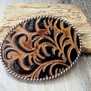 Puede incluir: Hebilla de cintur&oacute;n ovalada de cuero marr&oacute;n con un borde plateado estilo cuerda. La hebilla presenta un dise&ntilde;o floral en relieve detallado en marr&oacute;n oscuro sobre un fondo marr&oacute;n claro. Un accesorio de estilo western.