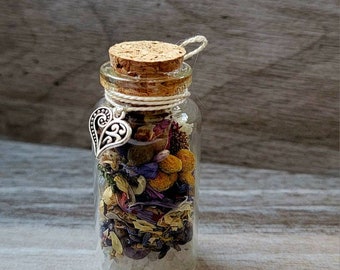 May include: A small glass bottle filled with dried flowers, herbs, and crystals. The bottle has a cork stopper secured with twine and a silver heart charm. The contents include yellow, purple, and white elements, creating a natural aesthetic.