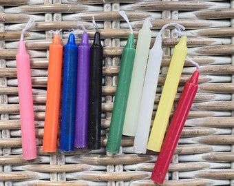 May include: A collection of nine colorful spell candles in shades of pink, orange, blue, purple, black, green, white, yellow, and red. Each candle is cylindrical with a white wick. The candles are arranged on a light brown woven surface.