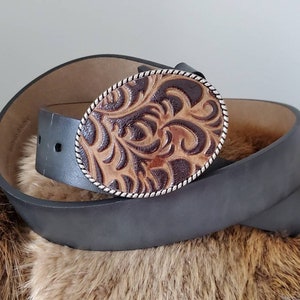 Puede incluir: Un cintur&oacute;n de cuero gris oscuro con una hebilla ovalada de cuero labrado marr&oacute;n y beige. La hebilla tiene un borde plateado y un dise&ntilde;o floral detallado. El cintur&oacute;n est&aacute; enrollado sobre una superficie marr&oacute;n texturizada.