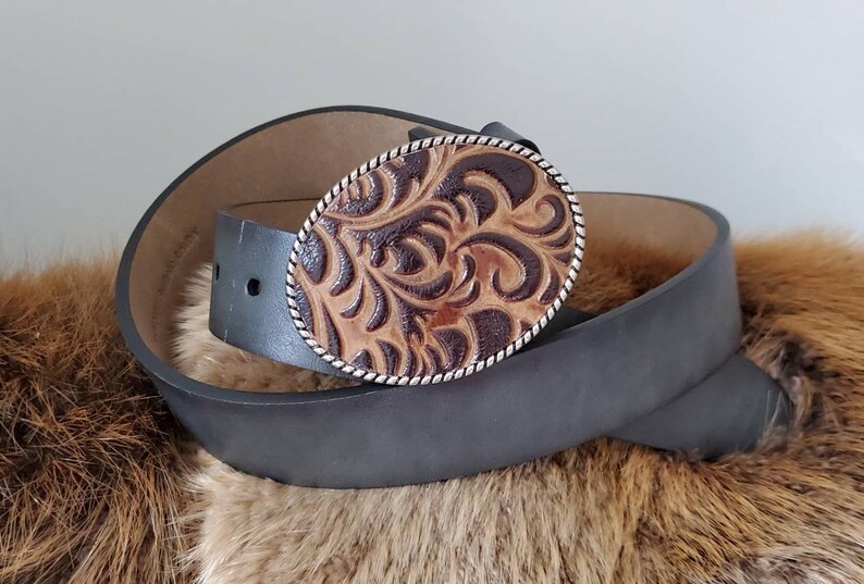 Puede incluir: Un cintur&oacute;n de cuero gris oscuro con una hebilla ovalada de cuero labrado marr&oacute;n y beige. La hebilla tiene un borde plateado y un dise&ntilde;o floral detallado. El cintur&oacute;n est&aacute; enrollado sobre una superficie marr&oacute;n texturizada.
