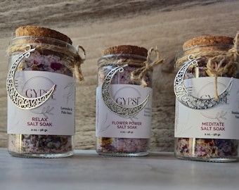 May include: Three glass jars of salt soak with cork lids and decorative silver crescent moon charms. Each jar has a label with the text "GYPSE" and the product name. The jars are filled with colorful bath salts and tied with twine.