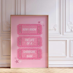 May include: A framed pink print featuring three ticket-shaped designs with the phrases "NOW I KNOW", "THE LIFE OF A", and "SHOWGIRL BABE" in white text. The print includes star accents and the number "511002".