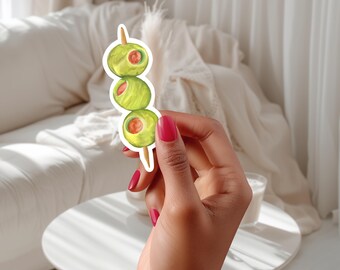 May include: A sticker of three green olives on a wooden skewer. The olives are drawn in a watercolor style with red centers.