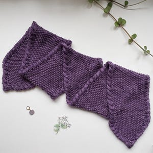 Può includere: Sciarpa lavorata a maglia in una tonalità viola intenso, piegata a zig-zag. La sciarpa presenta un punto a maglia strutturato e un dettaglio del bordo intrecciato. La sciarpa è esposta su una superficie bianca con un rametto di verde.