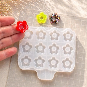 Puede incluir: Un molde transparente de silicona con forma de flor con 15 cavidades, junto a tres dijes de flores de resina acabados en rojo, verde y purpurina multicolor. El molde es para la elaboración de joyas y accesorios de resina.