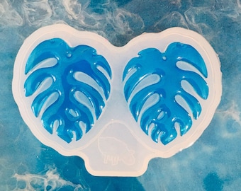 May include: A clear silicone mold with two leaf shapes. The leaves are in the shape of a monstera plant and are filled with a blue resin.