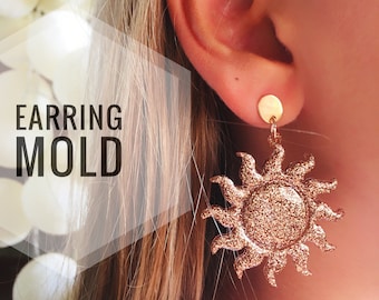 Könnte beinhalten: Ein Paar goldene Glitzer-Sonnenohrringe. Die Ohrringe werden von einer Person mit langen braunen Haaren getragen. Die Ohrringe sind ein beliebter Stil für den Sommer.
