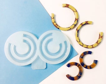 Può includere: Uno stampo in silicone per realizzare orecchini in resina. Lo stampo ha due cavità circolari, ciascuna con una forma curva per creare orecchini a cerchio. Nell'immagine sono presenti anche tre paia di orecchini in resina finiti, due paia con un motivo marrone e giallo e un paio con un motivo marrone e bianco.