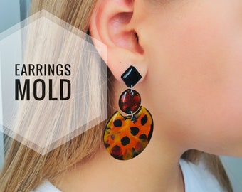 May include: A pair of dangle earrings with a black square-shaped top and a round, orange, black, and brown patterned bottom. The earrings are shown on a person's ear.