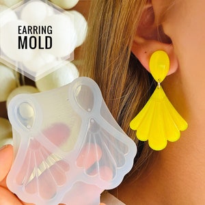 Può includere: Uno stampo in silicone trasparente per realizzare orecchini. Lo stampo ha due cavità, ciascuna a forma di ventaglio con una parte superiore a forma di lacrima. Lo stampo è mostrato accanto a un paio di orecchini gialli a forma di ventaglio.