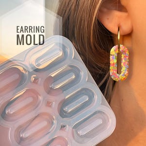 Puede incluir: Un molde transparente y flexible para pendientes con múltiples cavidades ovaladas. El texto "EARRING MOLD" está impreso en la imagen. Un pendiente de aro dorado con un colgante ovalado colorido y brillante se lleva en la oreja.