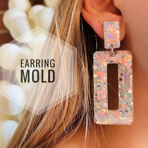 Peut inclure: Une paire de boucles d'oreilles rectangulaires transparentes avec une finition scintillante. Les boucles d'oreilles ont une petite découpe rectangulaire au centre. Les boucles d'oreilles sont de couleur rose clair et irisée.