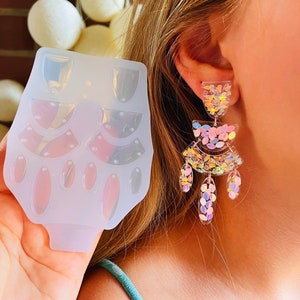 Puede incluir: Un par de pendientes de purpurina iridiscente con un diseño de varios niveles. Los pendientes están hechos de resina transparente y presentan una variedad de colores, incluyendo rosa, azul y amarillo. Los pendientes se muestran junto a un molde de silicona para hacer pendientes similares.