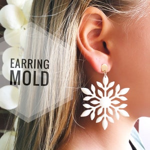 May include: White snowflake earring mold. The mold is made of a white material and is shaped like a snowflake. The mold is shown being worn by a person with blonde hair.