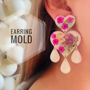 May include: A pair of white heart-shaped earrings with pink flowers and gold accents. The earrings have three teardrop-shaped dangles.