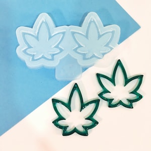 May include: A clear silicone mold with two cannabis leaf shapes, and two green cannabis leaf-shaped cutouts. The items are on a white and light blue background. The mold is for resin casting or similar crafts.