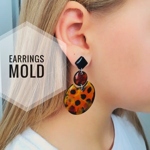 May include: A pair of dangle earrings with a black square-shaped top and a round, orange, black, and brown patterned bottom. The earrings are shown on a person's ear.
