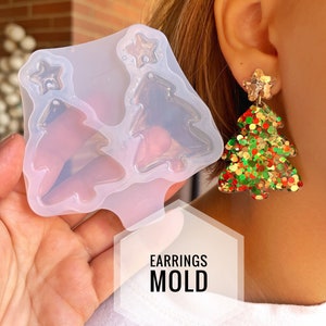 Puede incluir: Molde de silicona transparente para hacer pendientes de árbol de Navidad. El molde tiene dos cavidades para hacer pendientes. El molde se muestra junto a un par de pendientes terminados con purpurina roja, verde y dorada.