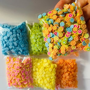 Puede incluir: Una colección de rodajas de fruta de arcilla polimérica de colores en bolsas de plástico transparente. Las rodajas presentan diseños de frutas naranjas, amarillas, rosas, verdes y azules. Las bolsas se sostienen en una mano, mostrando la variedad de colores y patrones.