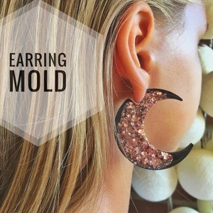 Puede incluir: Un par de pendientes en forma de media luna con contorno negro y relleno de purpurina rosa. Los pendientes se muestran en la oreja de una modelo.